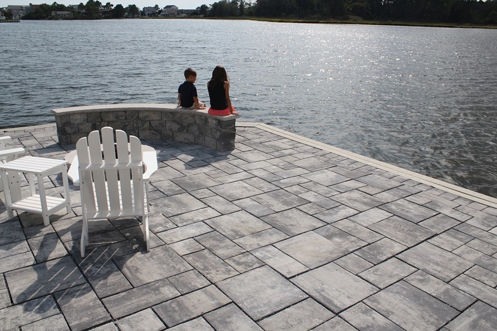 Two people sit on a stone bench by the water on a sunny day. The area is paved with gray tiles and features white Adirondack chairs. Trees and houses are visible in the distance across the water.