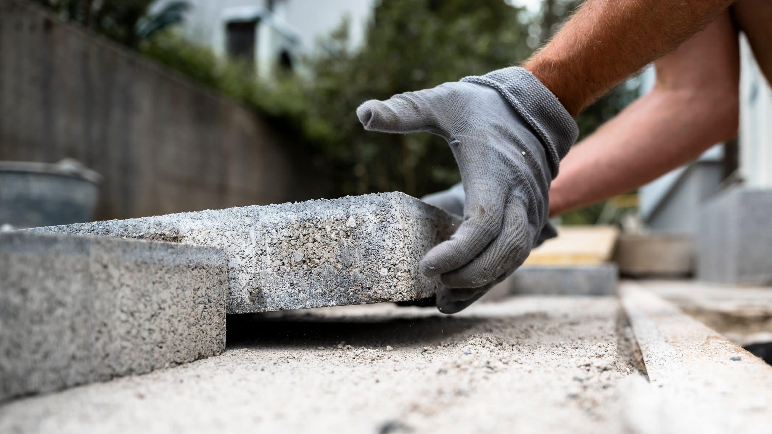 A person wearing gray work gloves is placing a concrete brick on the ground. The focus is on the hands and the brick, with other bricks laid in the background. The setting appears to be an outdoor construction site.