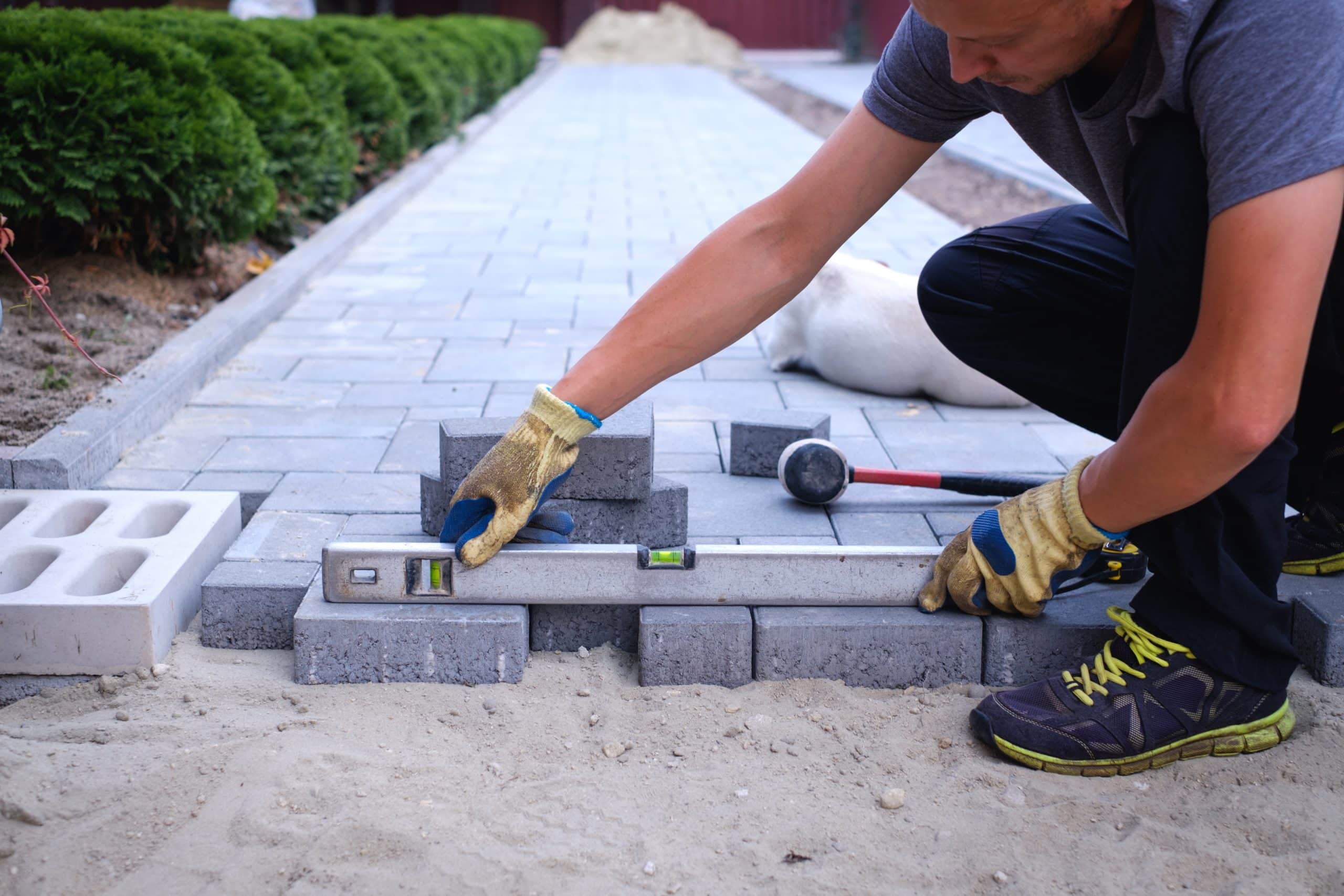 A person wearing gloves is laying gray concrete pavers on a sandy path. They are using a level to ensure the bricks are even. A rubber mallet and additional bricks are nearby. Shrubs line one side of the path.