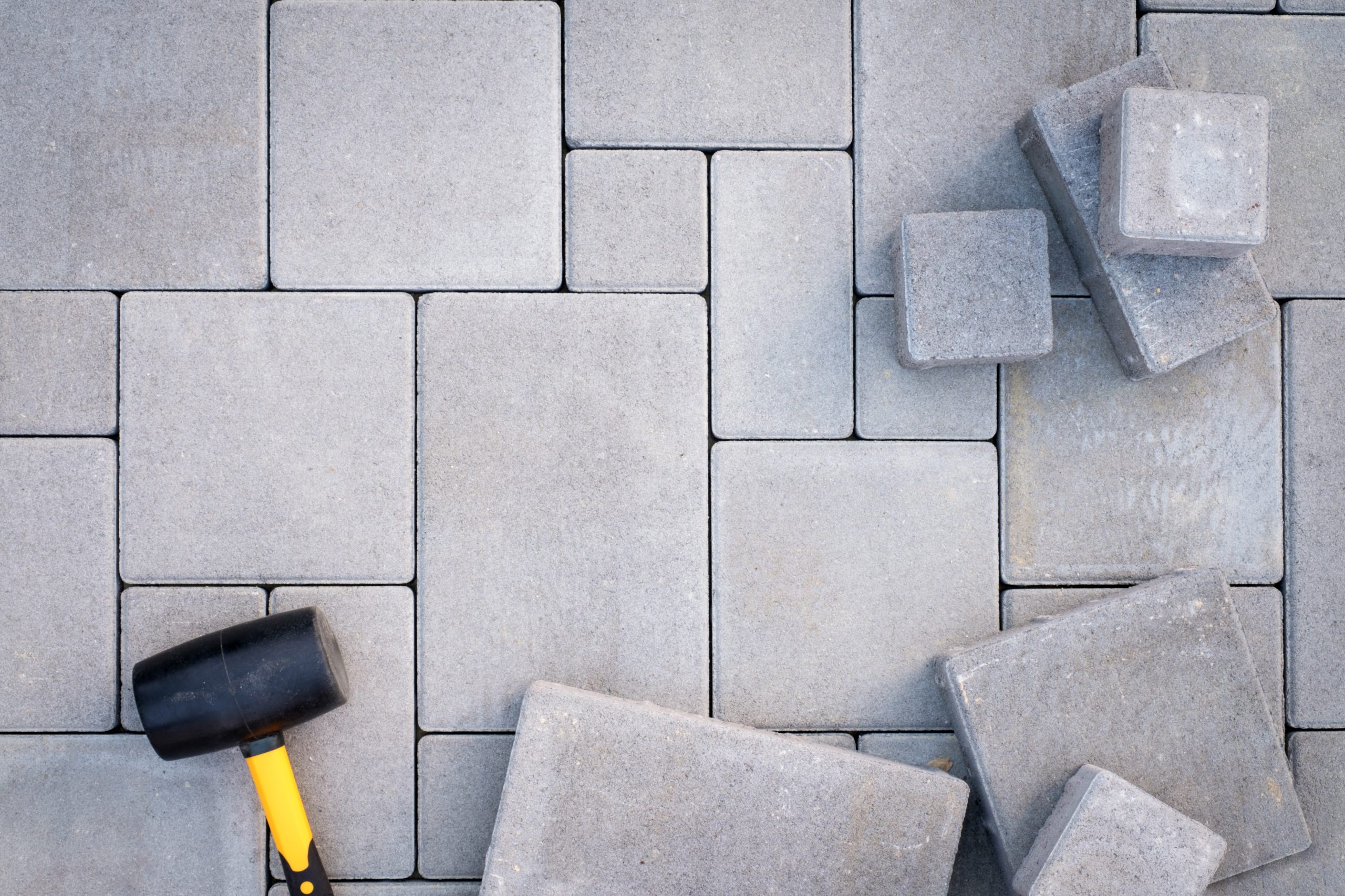 A partially completed stone pavement with interlocking gray bricks. A rubber mallet with a yellow handle lies on the surface, and a small pile of bricks is on the right side, suggesting ongoing work.