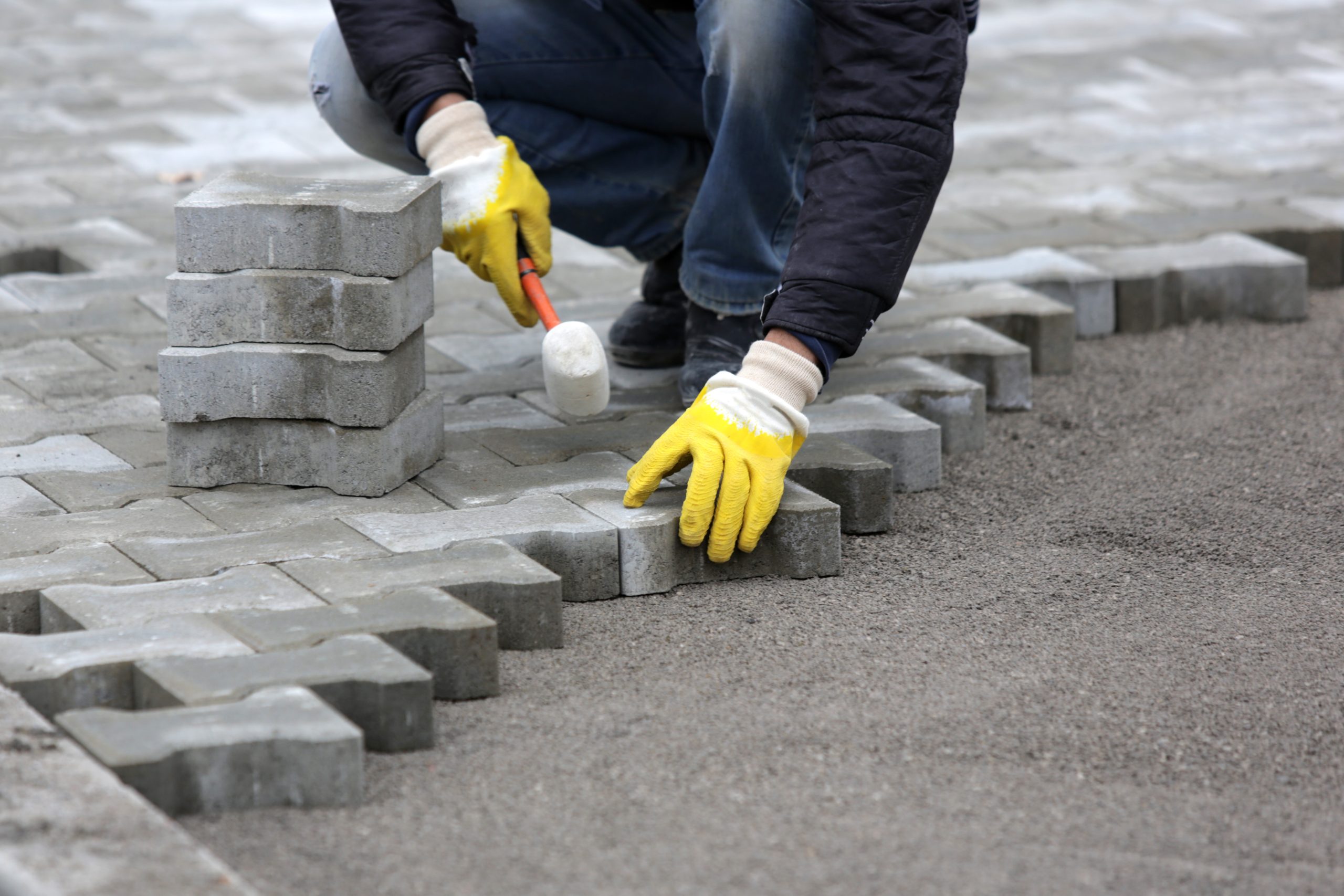 A person wearing yellow gloves is laying gray paving stones in a herringbone pattern on a sand and gravel base. They are using a rubber mallet to tap the stones into place, with stacks of stones visible nearby.