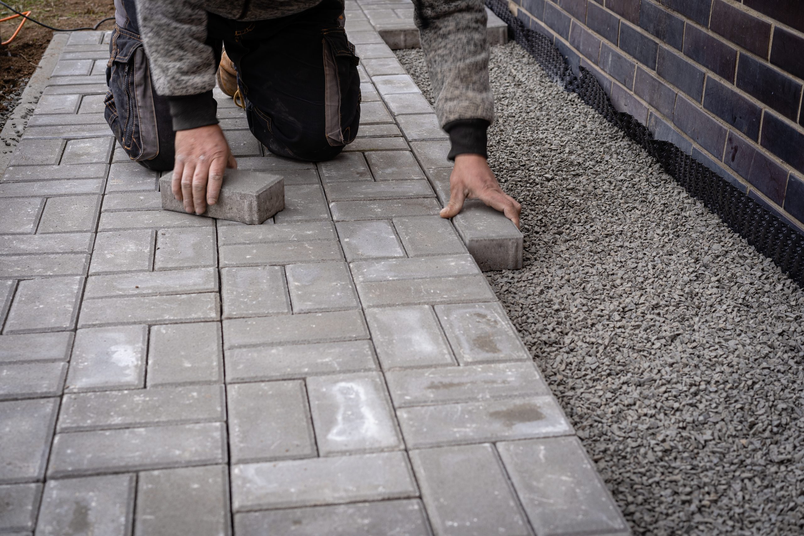 A person is kneeling and placing rectangular paving stones onto a gravel base next to a building. The paving stones are arranged in a staggered pattern. The person is wearing a gray sweater and work pants.