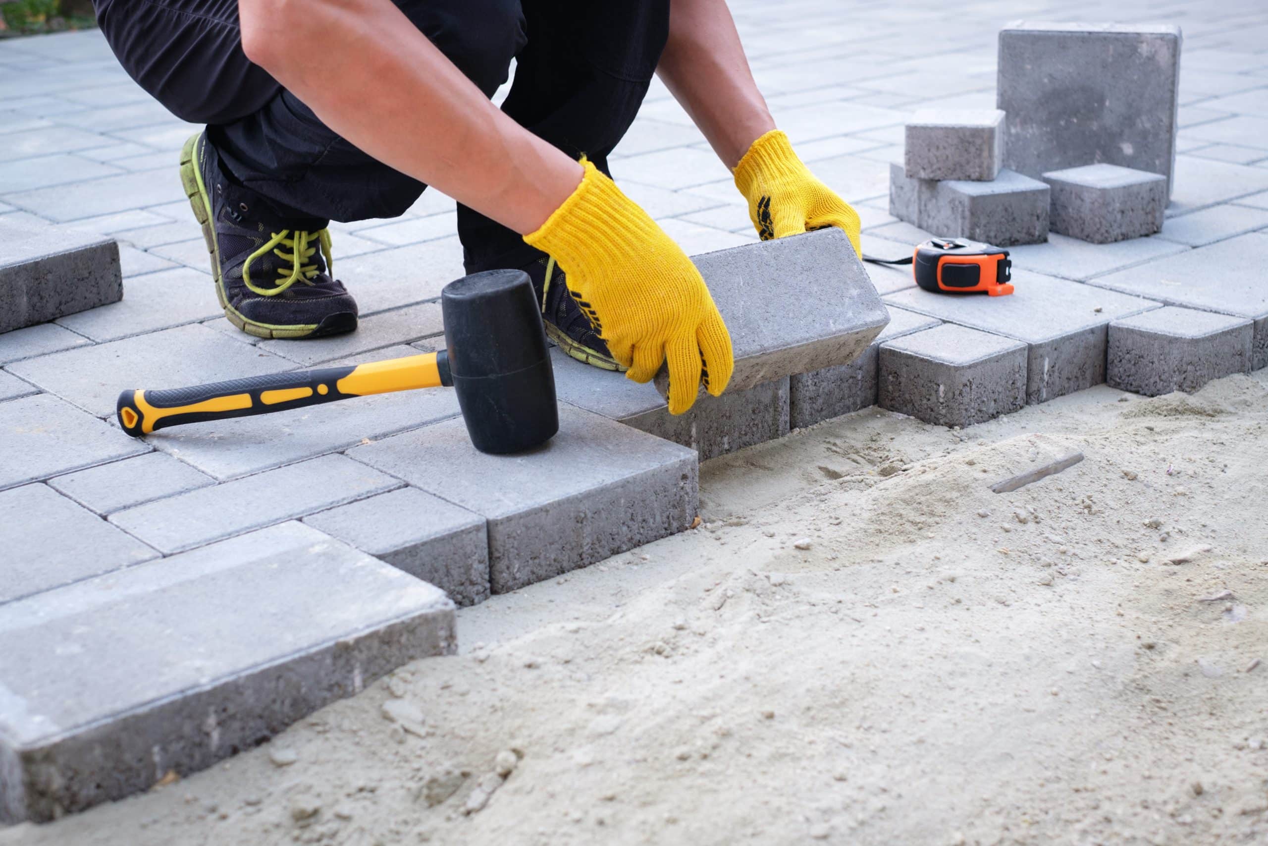 A person wearing yellow gloves is placing concrete pavers on a patio area. There is a rubber mallet and a tape measure beside the worker. The ground is partially covered with sand.