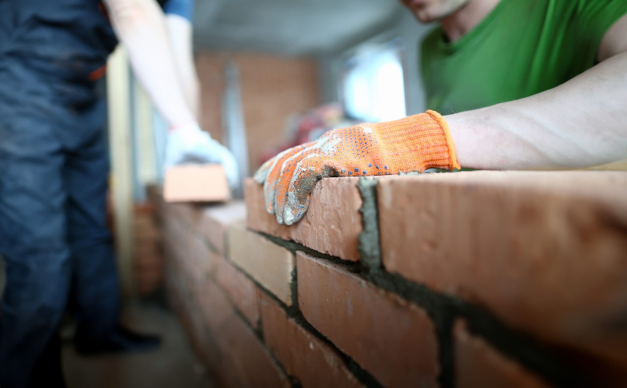 Two construction workers building a brick wall. One worker, wearing a green shirt and orange gloves, is placing a brick. The other worker in the background holds a brick, wearing blue overalls. The setting is indoors with unfinished walls.