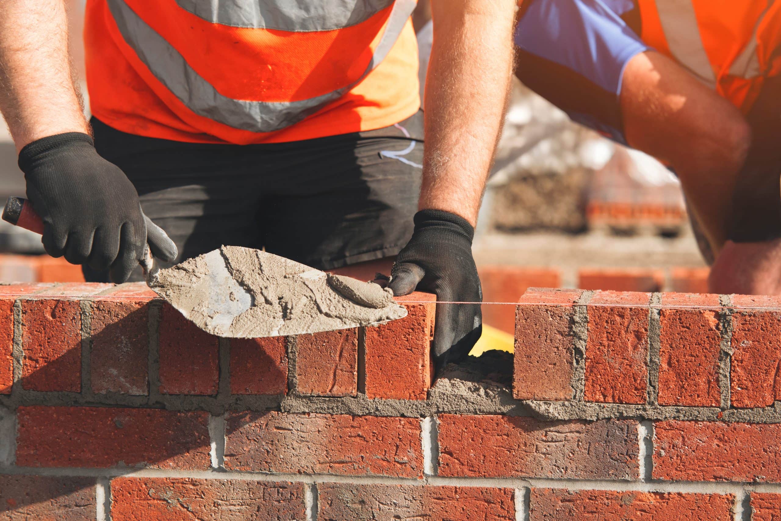 Two construction workers wearing orange vests and black gloves are building a brick wall. One is applying cement with a trowel, while a string line helps ensure a straight row of bricks. Sunlight illuminates the scene.