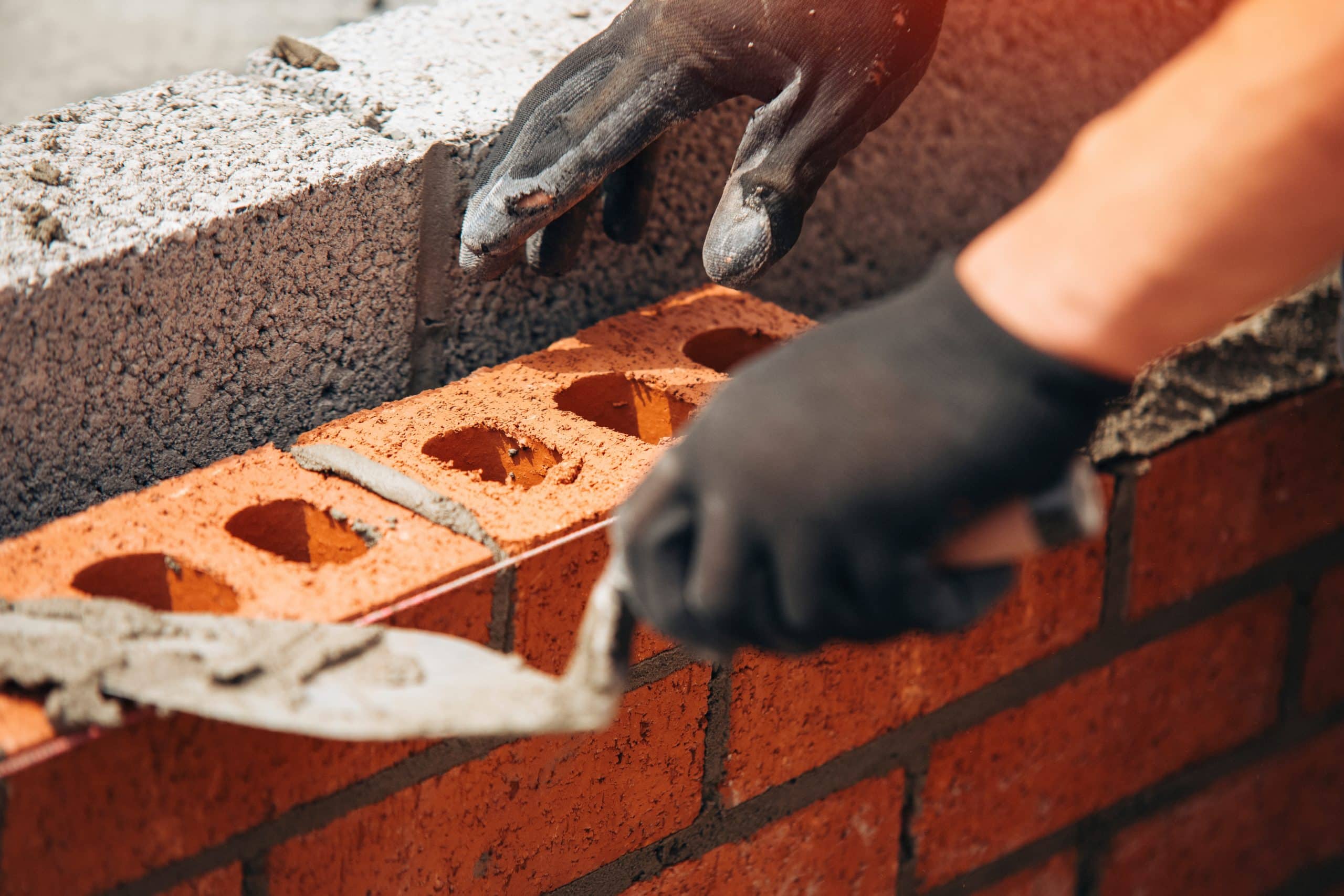 A person wearing black gloves is laying bricks and applying mortar with a trowel. The brick wall is partially constructed, showing a layer of red bricks with holes and another layer of gray concrete blocks in progress.