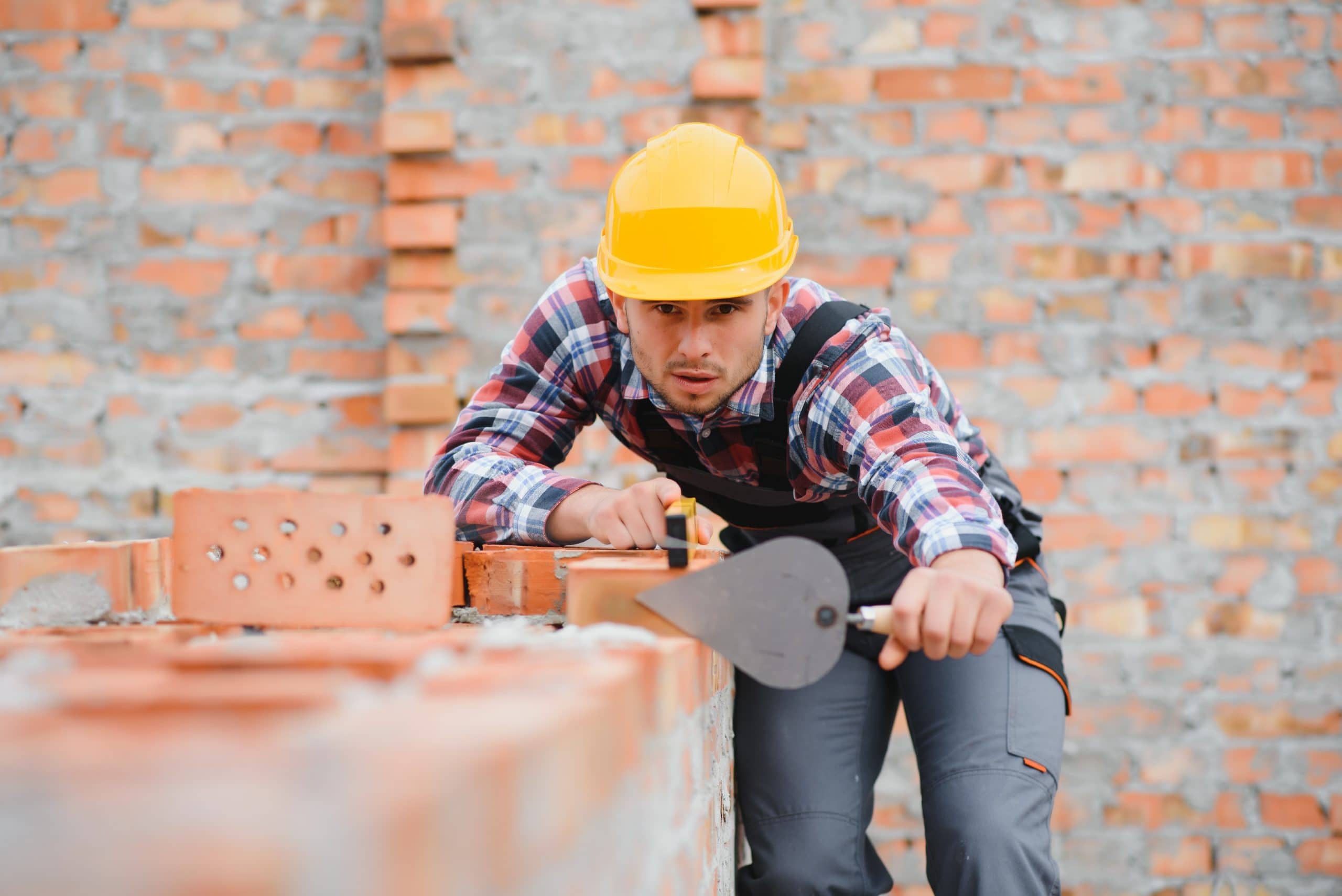 A construction worker wearing a yellow hard hat and plaid shirt is laying bricks. He is using a trowel to shape cement on a partially built brick wall, with a focus on precision. A brick wall forms the background.