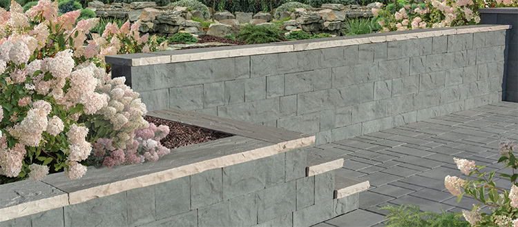 A stone retaining wall with light gray bricks runs alongside a pathway made of square stone tiles. The left side of the wall has blooming white hydrangeas and greenery. In the background, more foliage is visible.