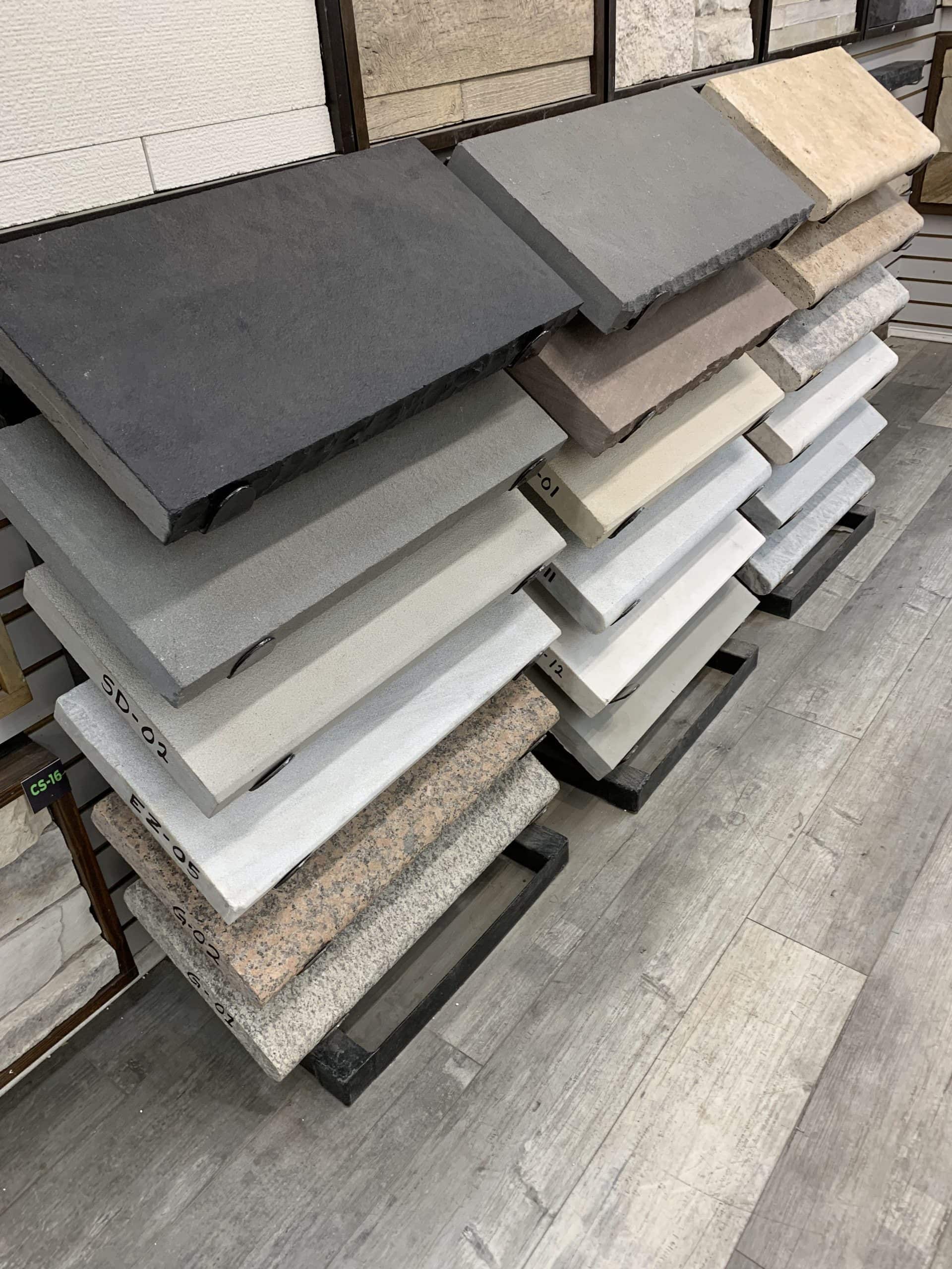 A display of various stone tiles in different shades, arranged vertically on a rack in a showroom. The tiles range from dark black and gray to beige, white, and speckled patterns, set against a wooden floor background.