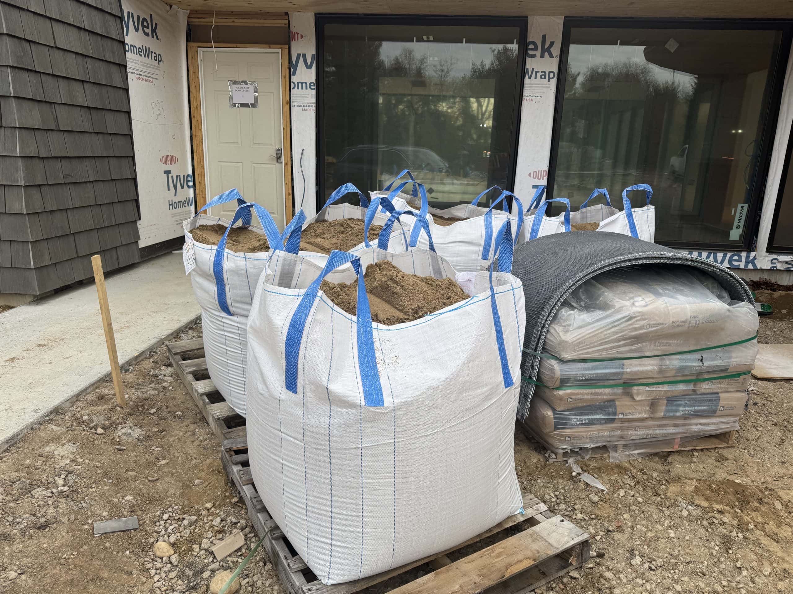 Large white bags filled with sand are placed on a wooden pallet at a construction site. Nearby are sheets of building material and covered equipment. The building in the background is partially wrapped in protective material.