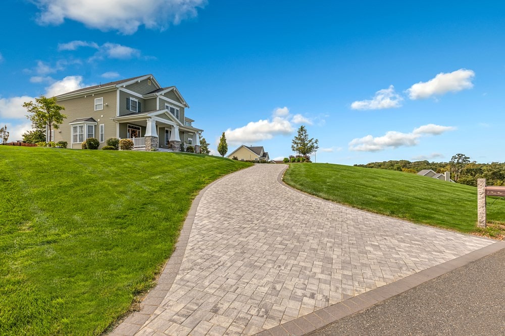 A two-story house with a gray exterior sits atop a lush green hill under a bright blue sky. A brick driveway curves up the hill leading to the house, with a few trees and shrubs lining the neatly manicured lawn.