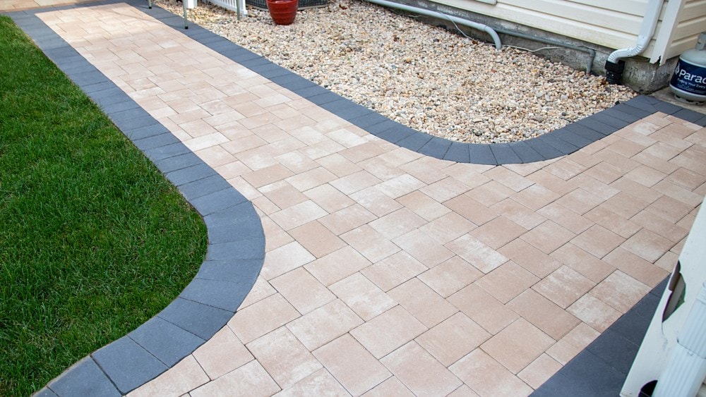 A neatly arranged brick pathway with beige and dark gray bricks curves between a grassy lawn and a gravel area next to a house. A potted plant and a drainage pipe are visible near the wall.