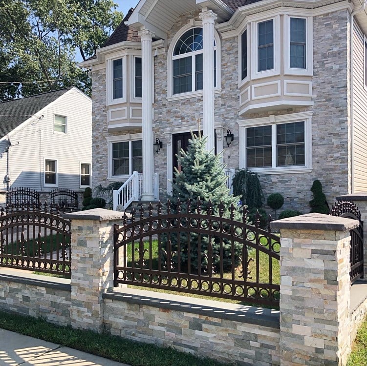 A two-story house with light stone exterior, tall white columns, and a manicured lawn featuring a small evergreen tree. The yard is enclosed by a decorative metal fence with stone pillars. Another house is visible in the background.