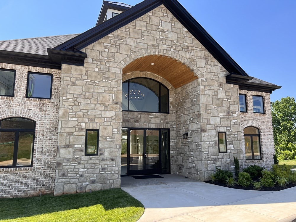 A stone and brick building with a large entrance. The entrance features a black-framed glass double door and a tall arched window. The facade includes a wooden arch ceiling, black trim, and surrounding greenery. Clear blue sky in the background.
