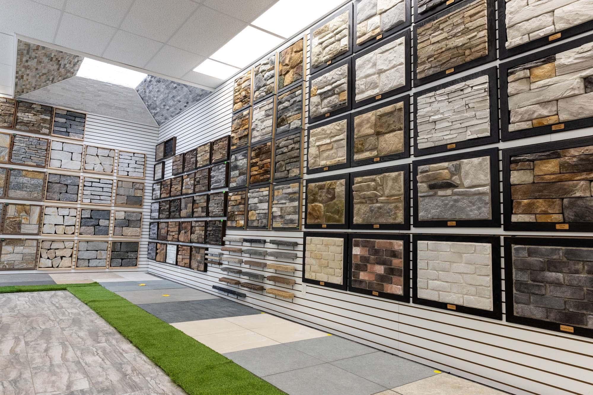 A showroom displaying various stone veneer samples on walls. The stones are arranged in square and rectangular panels, with different textures and colors. The floor has large tile samples and patches of green artificial grass.