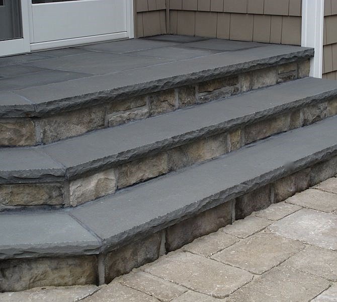 Stone steps leading up to a doorway, with light gray tiles on each step. The sides are made of a rough stone texture. The surrounding ground is paved with rectangular stones in a lighter shade.
