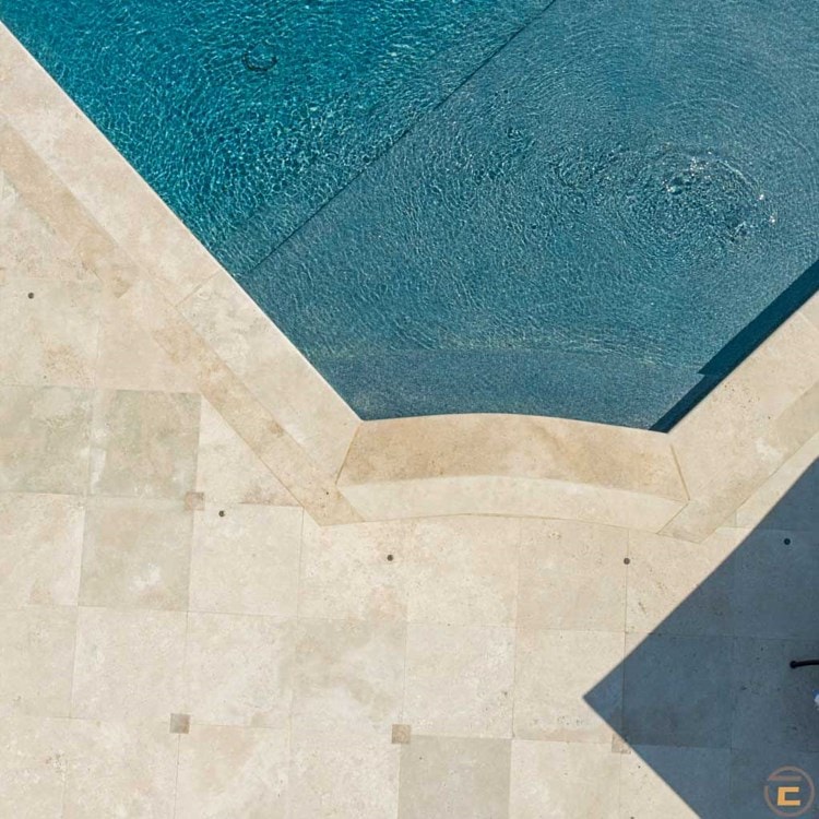Aerial view of a corner of a swimming pool with clear blue water. The pool is bordered by beige stone tiles, creating a geometric contrast. The corner design features a subtle curved edge and a small shaded area at the bottom right.