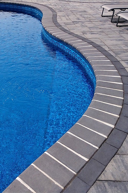 Curved swimming pool with clear blue water, surrounded by a paved stone deck. Two white lounge chairs are visible on the right side. The pool's edge is lined with gray bricks.