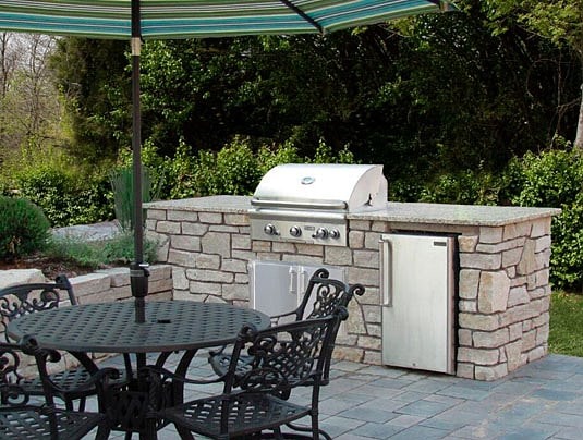 Outdoor kitchen area with a stone counter, stainless steel grill, and fridge. A round metal table with ornate chairs is shaded by a large striped umbrella. Lush greenery surrounds the patio, made of gray paving stones.