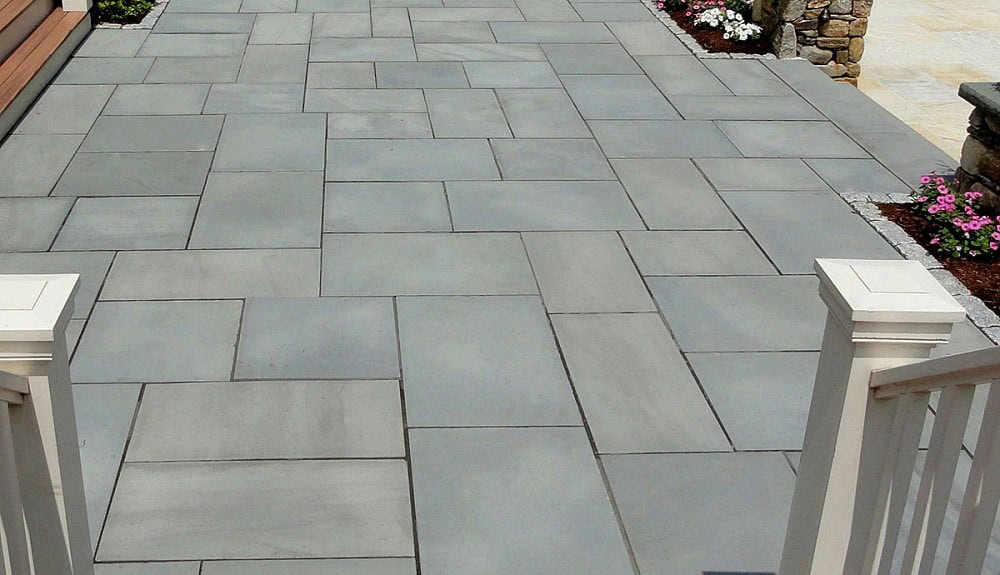 Large patio area paved with uniformly cut gray rectangular stones arranged in a geometric pattern. The area is surrounded by flower beds with pink and white flowers. White railings are in the foreground, leading up to the patio.