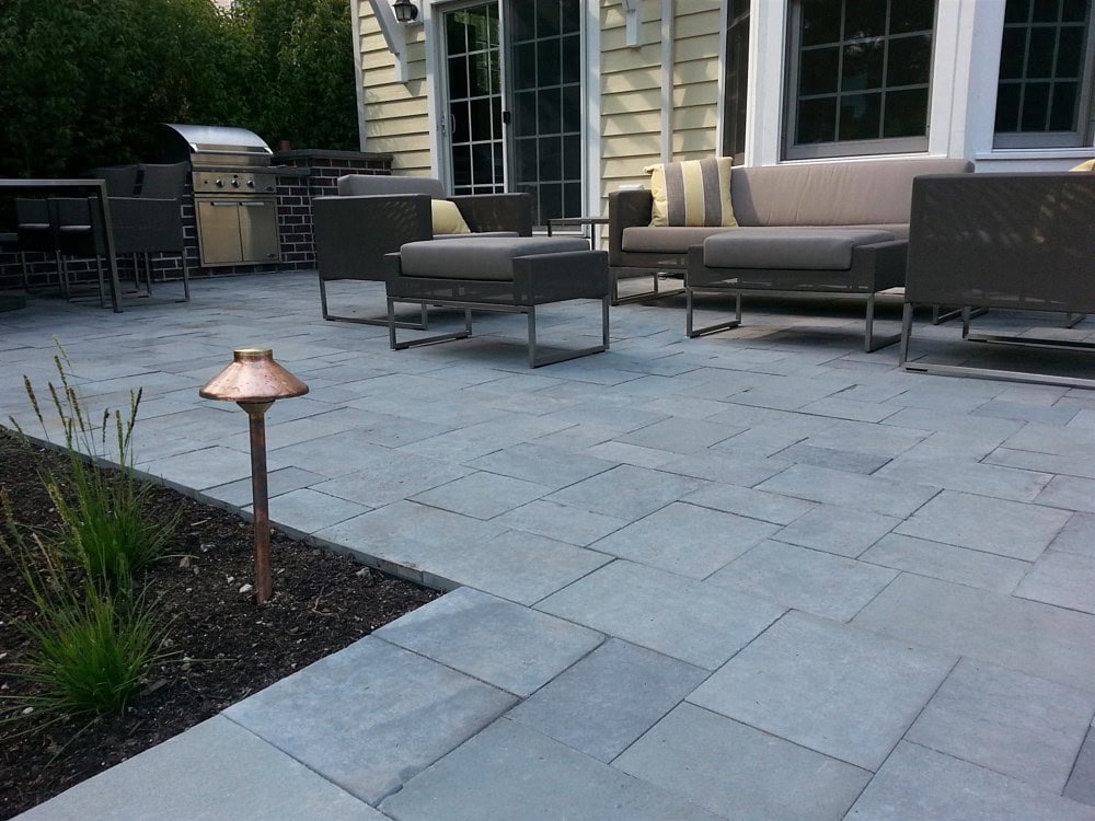 A modern patio with gray stone tiles features a seating area with a sofa and chairs, accented by cushions. A grill is nearby, positioned against a brick wall. A copper garden light is installed beside a small flower bed in the foreground.