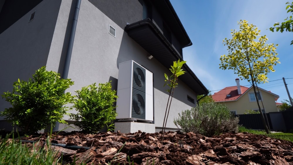 A heat pump unit is installed outside a modern house, surrounded by mulch, green plants, and young trees under a clear blue sky. Another house and trees are visible in the background.