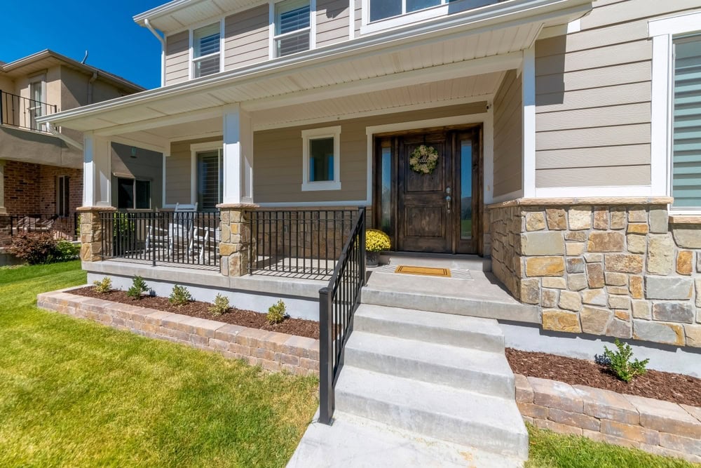 Front view of a modern two-story house with a beige exterior, stone accents, and a covered porch. A wooden front door has a wreath, and there are steps leading up to the entrance. A well-maintained lawn and small shrubs line the walkway.