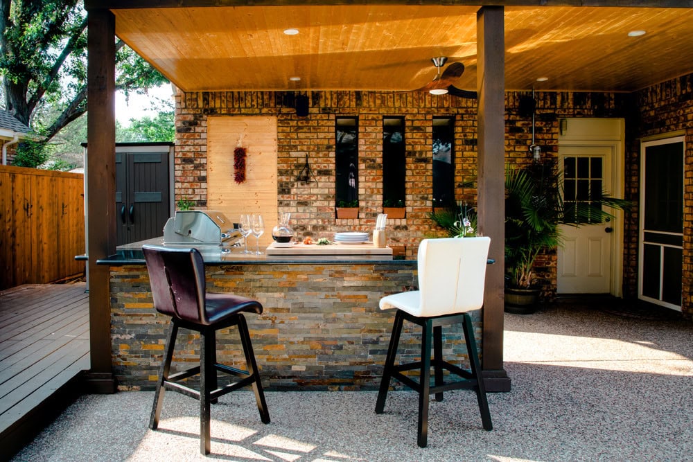 Outdoor kitchen with a stone island bar, two tall chairs (one black, one white), grill, and table settings. Brick walls, a wooden ceiling, and greenery create a cozy, stylish patio space.
