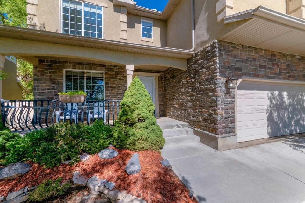 A modern townhouse exterior with tan stucco and stone accents, a white garage door, large windows, landscaped shrubs, a small tree, and a front porch with black railing and flower planter.