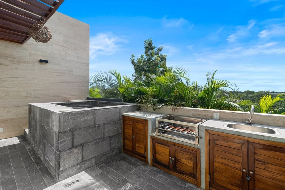 Outdoor kitchen with a stone Jacuzzi, a built-in grill with food cooking, wooden cabinets, and a sink, set against a backdrop of lush green plants and a clear blue sky.