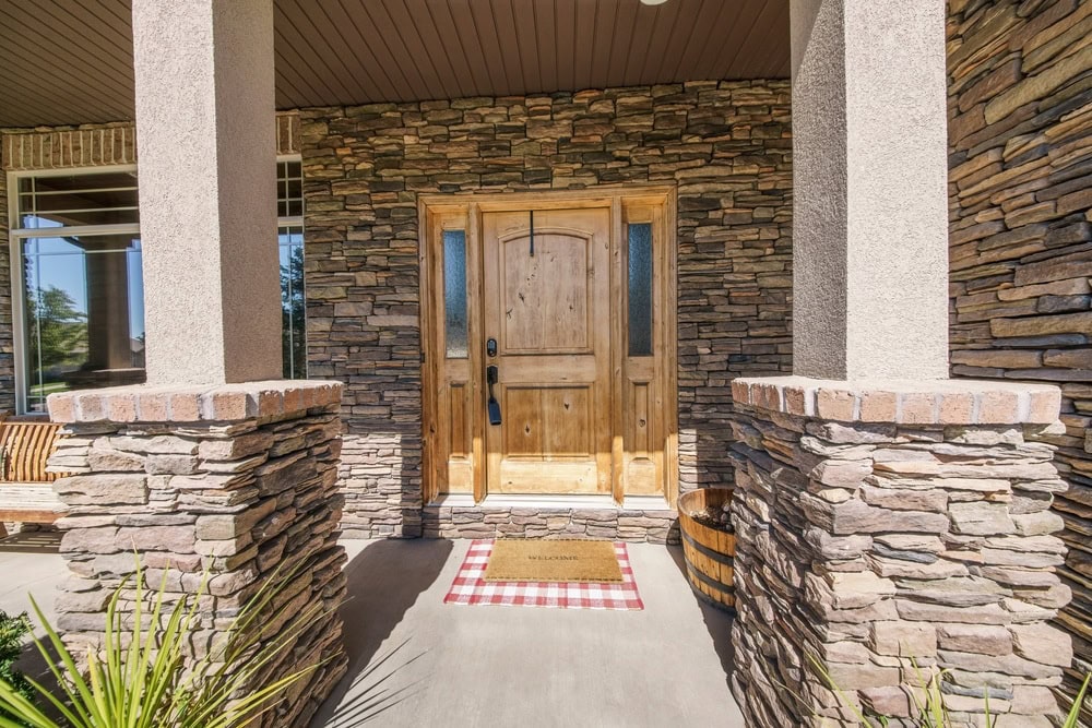 A wooden front door with decorative glass panels is set in a stone exterior. A red-and-white checkered rug and a welcome mat are in front of the entrance. Columns and a porch swing are also visible.