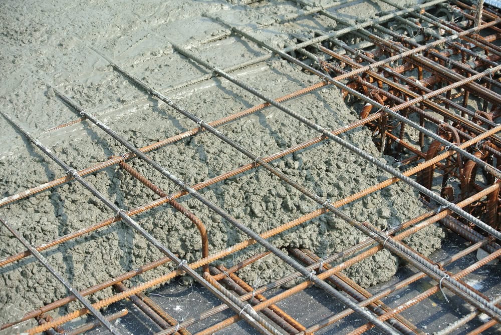 Close-up of wet cement being poured over a grid of rusted steel reinforcement bars at a construction site. The bars are arranged in a crisscross pattern, and the cement is spreading across them.