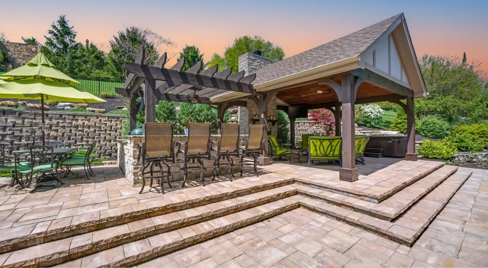 Outdoor patio with a covered seating area and bar stools under a pergola. A green umbrella shades nearby tables and chairs. The stone patio features steps and is surrounded by landscaped greenery with a sunset sky.