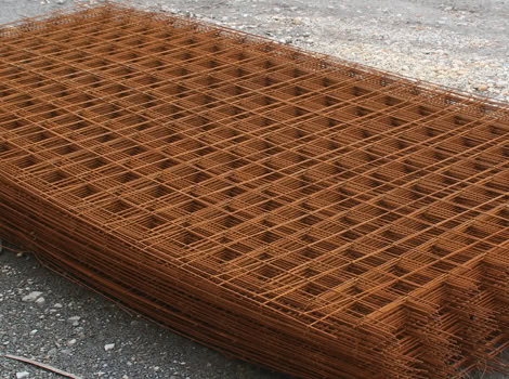 A large stack of rusty steel mesh sheets laid on bare ground. The mesh features a grid pattern with intersecting horizontal and vertical lines. Surrounding the stack are pebbles and gravel, indicating a construction or industrial setting.