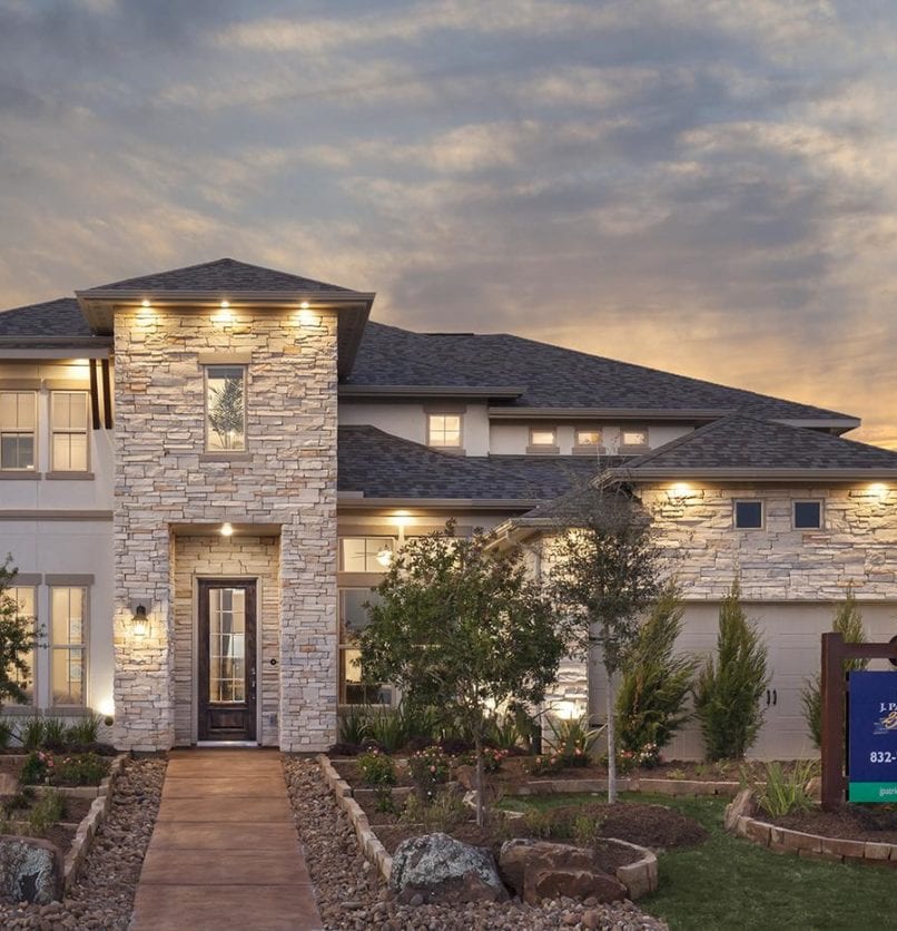 A modern two-story house with stone and stucco exterior, illuminated by warm lights at sunset, features large windows, landscaped front yard, and a sign near the entrance walkway.