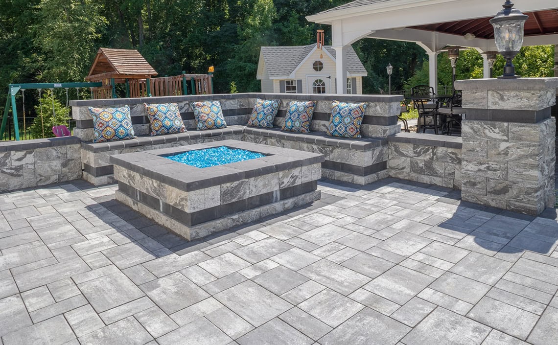 A modern outdoor patio with gray stone pavers, a built-in stone bench with colorful patterned pillows, and a square fire pit with blue glass. There’s a gazebo, playhouse, and swing set in the background.