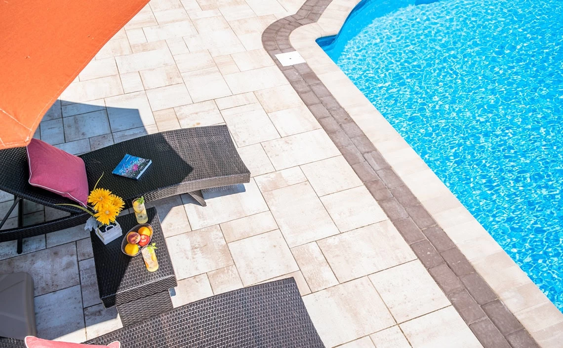 A bright, high-angle shot shows a sparkling blue swimming pool surrounded by light-colored patio pavers in Long Island