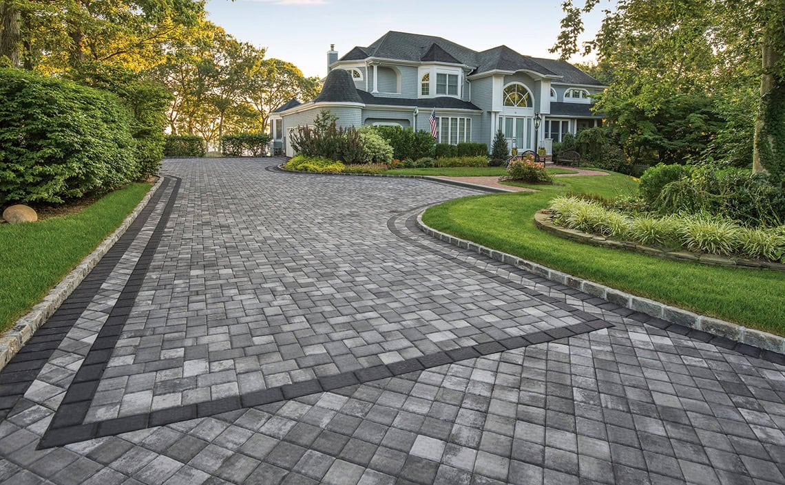 A large, elegant house with a distinctive roof sits at the end of a wide, winding driveway made of gray and black interlocking pavers, surrounded by manicured lawns and lush green landscaping.