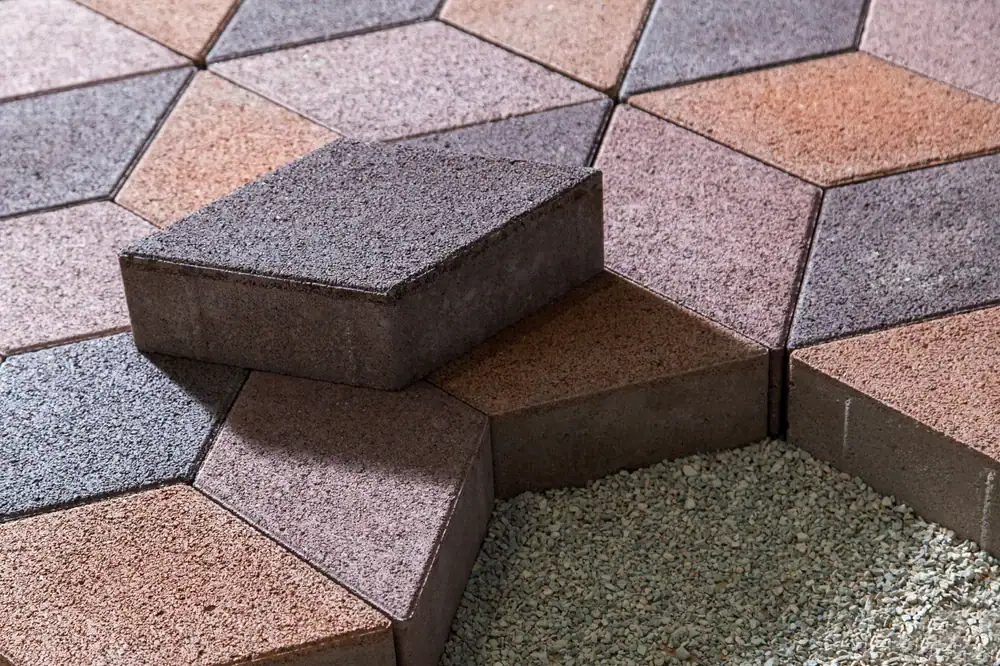 Hexagonal paving stones in shades of brown, pink, and gray are arranged in a geometric pattern. One stone is stacked on top of others, and some stones surround a patch of small gravel.