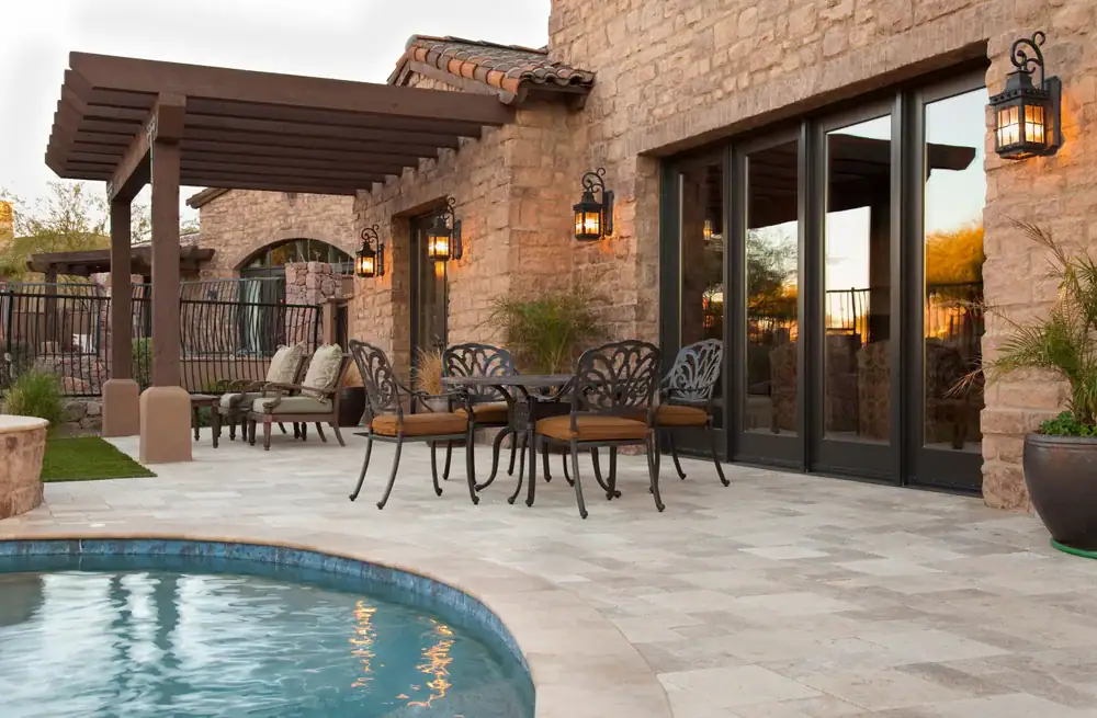 A stone patio features a round metal table with four chairs, a pergola with outdoor seating, potted plants, wall lanterns, and large glass doors, next to a curved swimming pool.